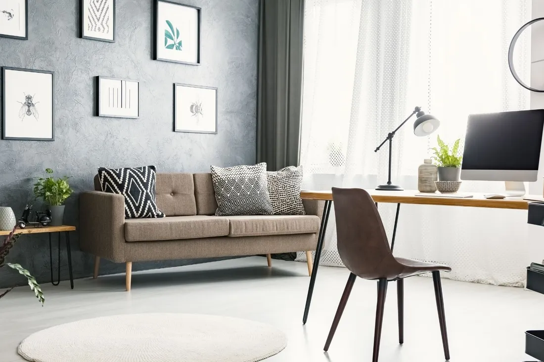 Kącik do pracy w salonie przy szarej ścianie z grafikami, beżowa sofa i zegar ścienny